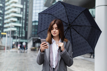 Happy businesswoman using mobile phone near office, young girl browsing phone smiling under umbrella while it is raining outdoors, female manager texting smartphone near modern bank building