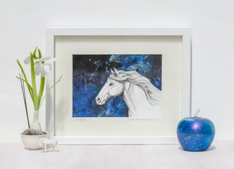 White interior display. Drawing of horses head on a collaged blue background in frame. With iridescent blue glass apple and snowdrop on a shelf.