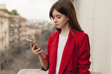 Attractive businesswoman using mobile phone standing on the balcony of office, young girl browsing phone smiling outdoors, portrait of female manager texting smartphone wearing red jacket and t-shirt