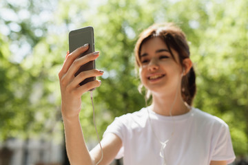 Happy girl smiling and laughing while having a video chat via mobile phone, pretty teenager using wireless connection on smart phone to make a video call sitting in the park, focus on hand with phone