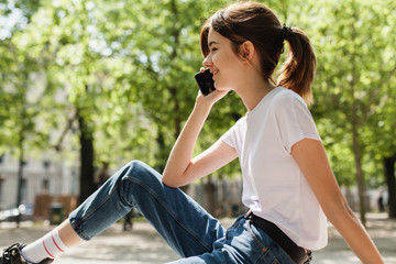 Hipster pretty student making a phone call while having a break in university and sitting on a branch in the park, happy girl talking via smartphone outdoors, smiling girl using cell phone for  a call