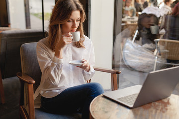 Young businesswoman drinking coffee while working on her laptop in coffee shop, beautiful female freelancer in casual style sitting relaxed at cafe near the window in armchair and watching video