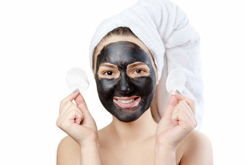  funny close-up portrait beautiful woman with facial black mask on white background, girl with a white towel on head, satisfied and happy  smile, holds in hands the cotton pads for removing make-up