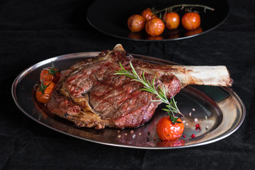 Single tomahawk rib steak on silver dish next to roasted cherry tomatoes