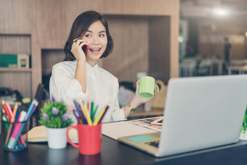 Happy asian woman working on desk talking on a cell phone with big smile and another hand holding a cup of coffee.