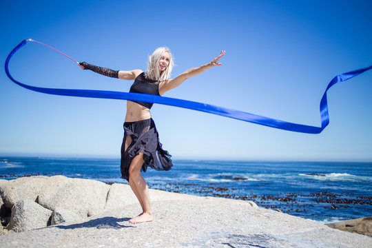 Beautiful female fitness model doing ribbon dancing on a granite rock overlooking the ocean on a bright and sunny summer day