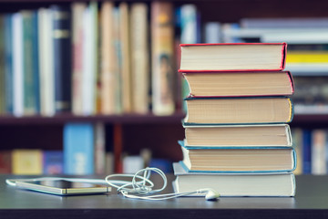 A stack of books and mobile phone with headphones on the background of bookshelves.
