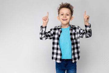 A handsome boy in a plaid shirt, blue shirt and jeans stands on a gray background. Boy smiling and showing thumbs up