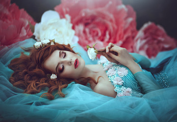Naklejka premium A beautiful sensual girl with red hair in a fairy blue lavish dress as a sleeping beauty lies in large flowers of pink and white peonies. The girl is a flower princess.