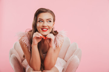 Portrait closeup of pleased smiling woman in dress propping her head while sitting in armchair, isolated over pink background