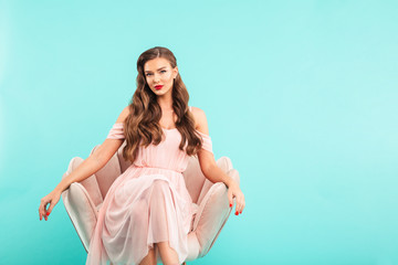 Image of gorgeous woman 20s in pink dress sitting in soft armchair with legs crossed and looking at...