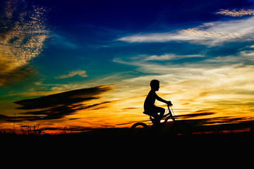 Silhouette of a boy riding a bicycle at sunset in public park.Orange blue sky on background.