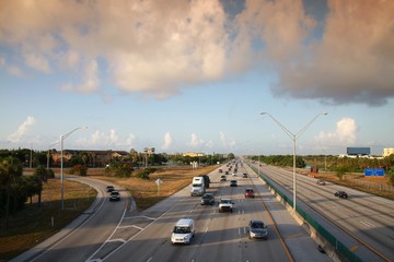 Traffic moves steadily along I-95 north and south on a hot sunny morning in rush hour under a cloudy sky.