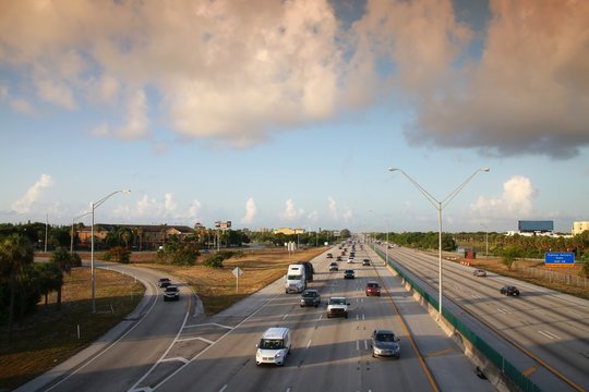 Traffic moves steadily along I-95 north and south on a hot sunny morning in rush hour under a cloudy sky.