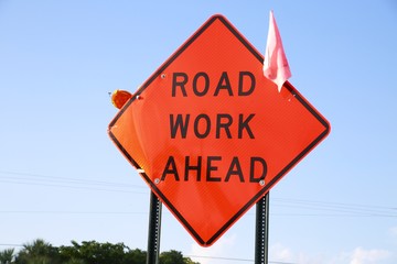 Orange Road Work Ahead Diamond Shaped Sign with Tiny Pink Flag, Clear Blue Sky and Trees Behind