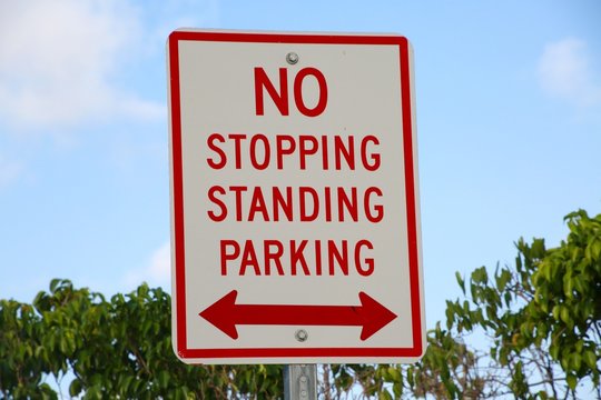 No Stopping Standing Parking Sign White Red Letters with Double-Arrows against Bushes Below