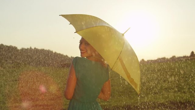 SLOW MOTION, CLOSE UP, LENS FLARE: Smiling blonde woman looks playfully over her shoulder and spins with her yellow umbrella on a refreshing rainy day in the summer. Happy girl dancing in spring rain.