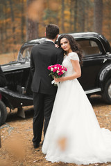Stylish wedding couple, bride, groom kissing and hugging near retro car in autumn