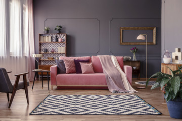 Pink sofa with two blankets and cushions standing in sitting room interior with windows with curtains, patterned carpet and empty frame on the wall