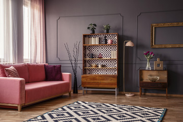 Retro wooden cupboard with books and decor standing in dark room interior with pink sofa, carpet and flowers in glass vase
