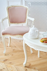 White wooden round table with tea pot, croissant and macaroons in white plate and pink soft chair near. Beautiful modern interior, copy space