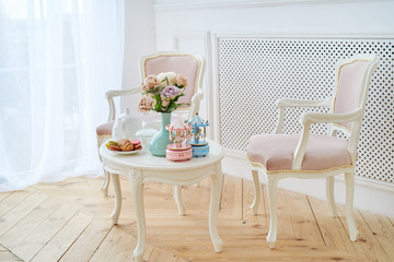 Two pink soft chairs and white wooden round table with breakfast, vase with flowers and merry-go-round musical carousel with horses. Beautiful modern interior, copy space