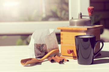 hot drink on sunny day/ black mug with cinnamon sticks and coffee beans is on the table on the background of hand grinder