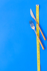 Diet, weight loss, slimming concept. Fork and knife with wound measuring tape on blue background...