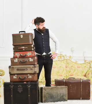 Macho elegant on tired face, exhausted at end of packing, leans on pile of vintage suitcases. Man with beard and mustache packed luggage, white interior background. Baggage and relocation concept.