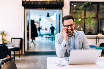 Smiling man talking over the phone while sitting in front of a laptop. Looking at camera.