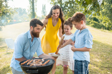 Dedicated father helping his son to use wooden tongs for putting sausages on the charcoal barbecue grill outdoors during family picnic in summer