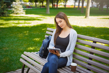 Motivated asian freelancer in the park. Girl on a bench, drinking coffee and working with tablet