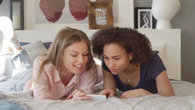 Close-up of two young attractive women caucasian blonde and watch scroll and discuss app on phone in a cozy bed in an airy sunny room in Scandinavian style design with pastel coloured textile