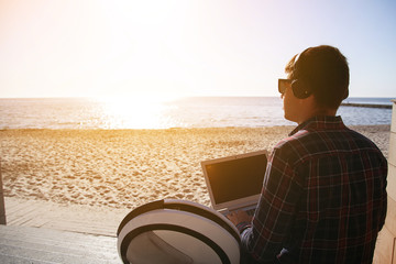 Fototapeta premium The guy at dawn is working in the laptop. Right next to it is a monoclean. The headphones are on. Looks at the sea, sitting on the beach.
