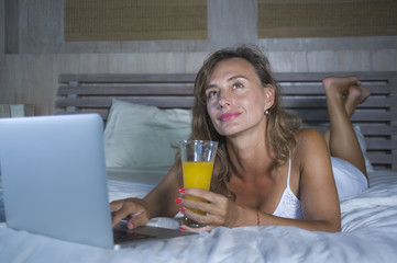 beautiful Caucasian woman 30s lying on bed at night in home bedroom using internet at laptop computer drinking healthy orange juice