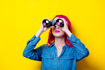 Young pink hair girl in blue shirt holding a binoculars. Portrait on isolated yellow background
