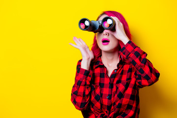 Young redhead girl with binoculars. Portrait isolated on yellow background