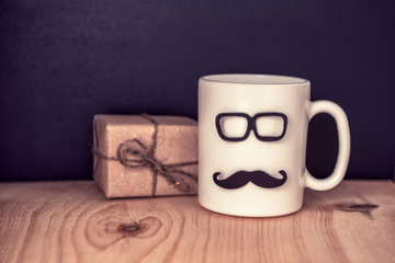 cup with mustache, glasses and gift box over chalkboard on wooden background, holiday concept of Fathers Day