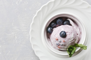 Close-up of blueberry ice cream and blueberries with mint leaves in a white bowl on a gray background.