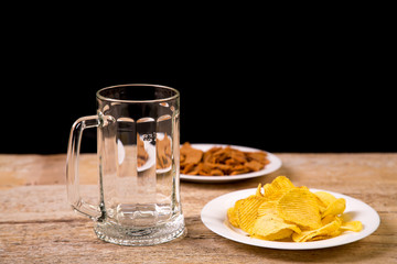 On a wooden table is a mug for beer. Nearby is a white plate with potato chips. Horizontal photo.
