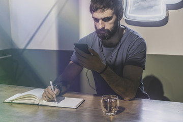 Young bearded man sitting in cafe and using mobile phone for listening digital music. Man making notes at note book. Horizontal. Blurred background