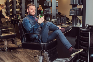 Fototapeta na wymiar A professional hairdresser sitting on an armchair, has a rest after work with a beer bottle in his hand.