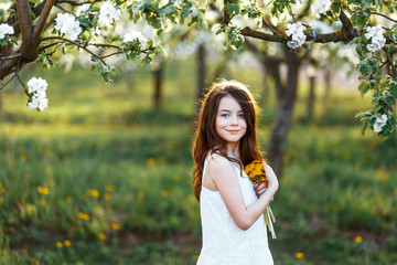 A portrait of a beautiful young girl with blue eyes in a white dress  in the garden with apple trees blosoming having fun and enjoying smell of flowering spring garden at the sunset