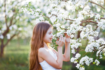 A portrait of a beautiful young girl with blue eyes in a white dress  in the garden with apple trees blosoming having fun and enjoying smell of flowering spring garden at the sunset