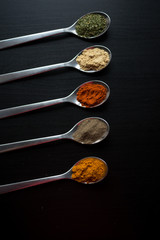 Spices colored in silver spoons, placed on a black wooden table. Pepper, pepper, salt, basil, mustard