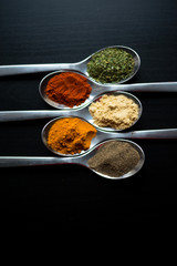 Spices colored in silver spoons, placed on a black wooden table. Pepper, pepper, salt, basil, mustard