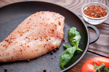 fresh raw chicken fillet sprinkled with herbs and spices and a basil leaf on a black frying pan on a wooden table with tomatoes and pepper in a container