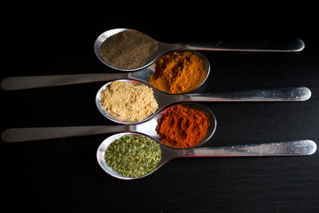 Spices colored in silver spoons, placed on a black wooden table. Pepper, pepper, salt, basil, mustard