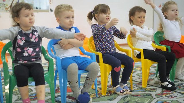 Children's developing a game room. Emotions of young children during entertaining classes. Children sit on chairs in a row. The child embraces himself with his hands and then raises them to the side.
