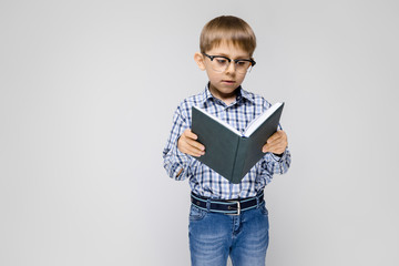 A charming boy with an inlaid shirt and light jeans stands on a gray background. The boy is holding a book in his hands. Boy with glasses